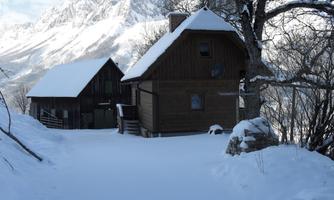 Two wooden buildings in the snow, surrounded by mountains. The sky is clear and the landscape looks peaceful.