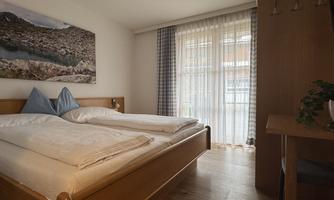 A cozy bedroom with a double bed and a large window front. The walls are light-colored and a picture hangs above the bed.