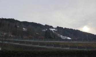 A wooded slope with snow-covered mountain slopes. The sky is cloudy and it is slightly twilight.