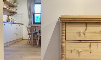 A cozy room with a wooden cupboard in the foreground. In the background, there is a bright kitchen with a table and chairs.