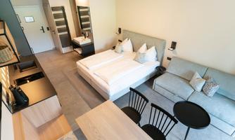 A modern hotel room with a double bed, a seating area, and a dining table. The decor is clean and stylish, with neutral colors and plenty of natural light.