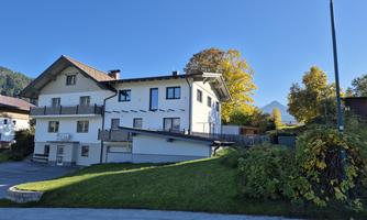 A modern house with a white facade and a green entrance. In the background, mountains and autumn trees can be seen.
