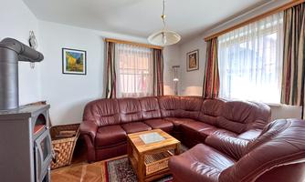 A cozy living room with a large, curved leather couch and a wooden table in the center. The walls are decorated with windows and pictures.