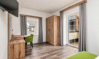 A modern room with wooden floors and large windows. There is a desk, a TV, and a wardrobe.