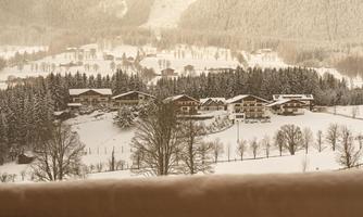 A snow-covered landscape with mountains in the background. In the foreground, houses and trees are visible, surrounded by a peaceful winter atmosphere.