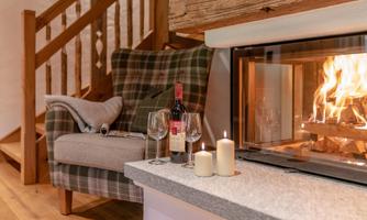 A cozy room with a comfortable armchair next to a fireplace. On the table are wine glasses, a bottle of wine, and candles.