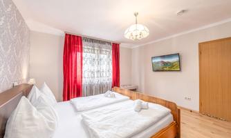 A cozy bedroom with a double bed and white bedding. The room has a warm atmosphere with red curtains and a TV on the wall.
