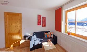A cozy living room with a blue sofa, colorful cushions, and a wooden table. The window offers a view of the mountains and lets in plenty of light.