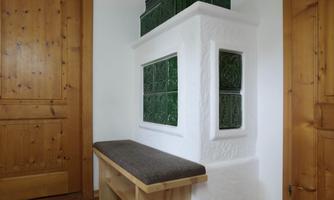 A traditional tiled stove with green ceramic and a wooden bench. The walls are made of wood, creating a cozy atmosphere in the room.