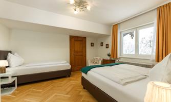 A bright room with two beds and a cozy window seat. The walls are painted in neutral colors, and there are wooden details in the room.