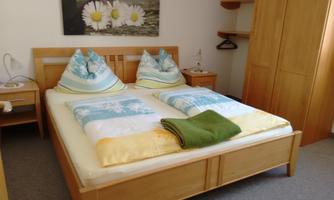 A cozy bedroom with a large double bed and colorful cushions. A picture of daisies hangs on the wall, and there is a wardrobe as well as bedside tables.