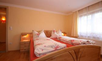 A cozy bedroom with two beds and colorful bedcovers. The room has bright walls and plenty of daylight coming through the large window.