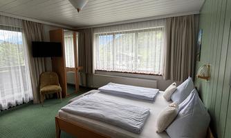 A cozy bedroom with a double bed and bright curtains. A television is mounted on the wall and a lot of natural light streams through the windows.