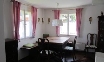 A cozy dining area with a wooden table and chairs. Bright walls and windows with red curtains create an inviting atmosphere.