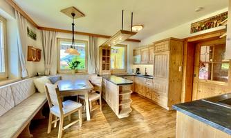 A modern kitchen with wooden shelves and a dining table. Large windows provide plenty of daylight and a view of the outdoors.