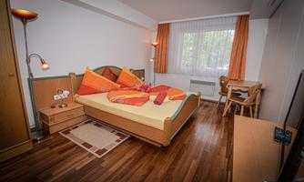 A cozy room with a double bed, colorful cushions, and wooden flooring. There is a small workspace and a window with a view.