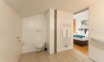 A modern bathroom with bright tiles and a towel warmer. In the background, a bedroom is visible.