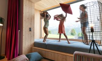 Three children are playing with pillows in a modern, bright room. The landscape is visible through the large window.