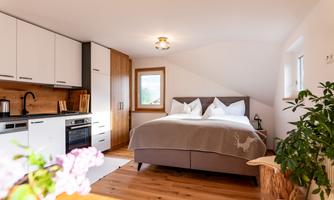 A modern bedroom with a large bed and bright pillows. The kitchen is open and well-equipped, surrounded by warm wooden tones.