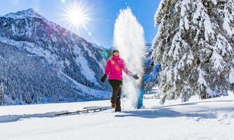 A woman in pink clothing happily walks through the snow. In the background, there are snow-covered mountains and bright sunshine.