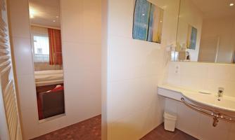 A modern bathroom with a sink, mirror, and a bright, minimalist design. In the background, a bed in a bedroom is visible.