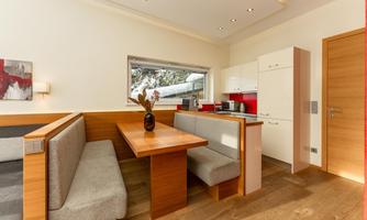 A modern dining room with a wooden tabletop and cozy benches. The kitchen in the background is bright and inviting.