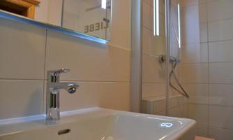 A modern bathroom with a simple sink and a shower. The walls are designed with light tiles, and there is a large mirror.