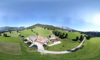 A picturesque landscape with a green meadow and a farm. In the background, mountains and a clear sky can be seen.