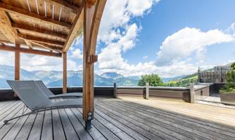 A cozy terrace with wooden flooring and modern loungers. In the background, the mountains and a clear sky are visible.