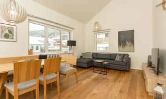 A modern living room with a dining table and chairs. Large windows offer a view of the surrounding landscape.