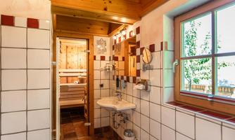 A modern bathroom with white tiles and wooden details. In the background, a sauna is visible and the window lets in plenty of daylight.
