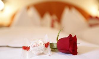 A red rose petal lies next to a candy on a bed. The atmosphere is romantic and inviting.