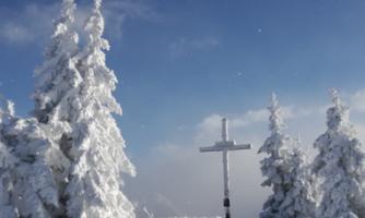 A snow-covered mountain peak with a white cross figure and snow-covered trees. The sky is clear and blue.