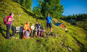 A group of hikers is taking a break in nature. In the background, a wooden house and green hills can be seen.