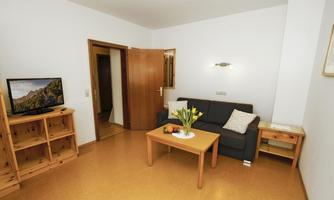 A bright living space with a comfortable sofa and a table. On the wall is a television and there is an open access to another room.