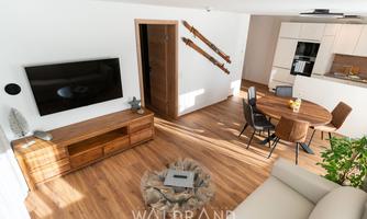 A modern living room with wooden flooring, television, and a round dining table. The decor is bright and friendly, with plants for a cozy atmosphere.
