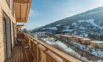 A balcony with a wooden railing overlooking a snow-covered landscape. In the background, there are mountains and a town with colorful buildings.