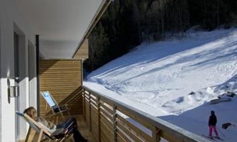 A woman is relaxing on a balcony with a view of a snow-covered landscape. In the background, a skier can be seen.