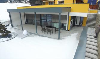 A modern house with a covered terrace and a table with chairs. The area is surrounded by snow, and there are stairs on the right side.
