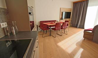 A modern living kitchen with a dining table and red chairs. The room has large windows and bright wooden floors.
