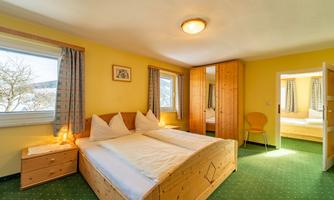 A cozy bedroom with two beds and yellow walls. The room has large windows and a wooden wardrobe.