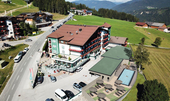 A hotel building in a picturesque mountain landscape, surrounded by green meadows. Nearby are parking spaces and a terrace with loungers.