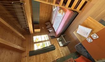 A cozy wooden room with a double bed and a small table. There is a wooden ladder on the wall that leads to a sleeping area.
