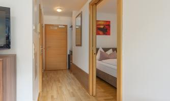 A bright hallway with wooden flooring and modern walls. In the background, there is a bedroom with a bed and a red artwork.