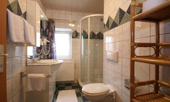 A modern bathroom with a shower, sink, and toilet. The walls are tiled, and a wooden shelf stands in the room.