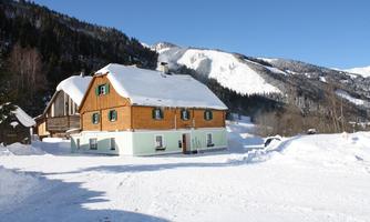 An idyllic wooden house in the snow, surrounded by mountains. The sky is clear and blue, creating a peaceful winter landscape.