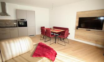 A modern living room with a cozy couch, a dining table, and red chairs. There is a television on the wall, and the kitchen is well-equipped.