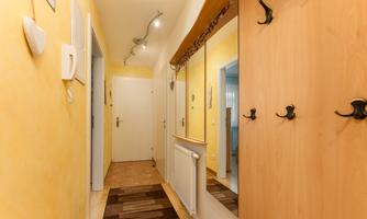 A bright hallway with yellow walls and a mirror. On the left are coat hooks and a door that leads to another room.