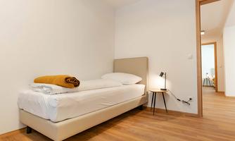 A simple bedroom with a single bed and a nightstand. There is a wooden floor and plenty of natural light.