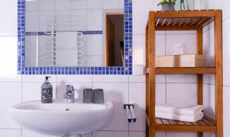 A modern bathroom with a sink and a large mirror. In the background, there is a wooden shelf with towels and toiletries.